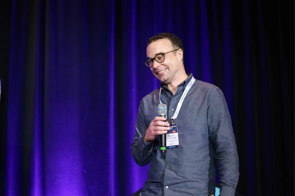 a man on stage, smiling as he presents, holding a microphone