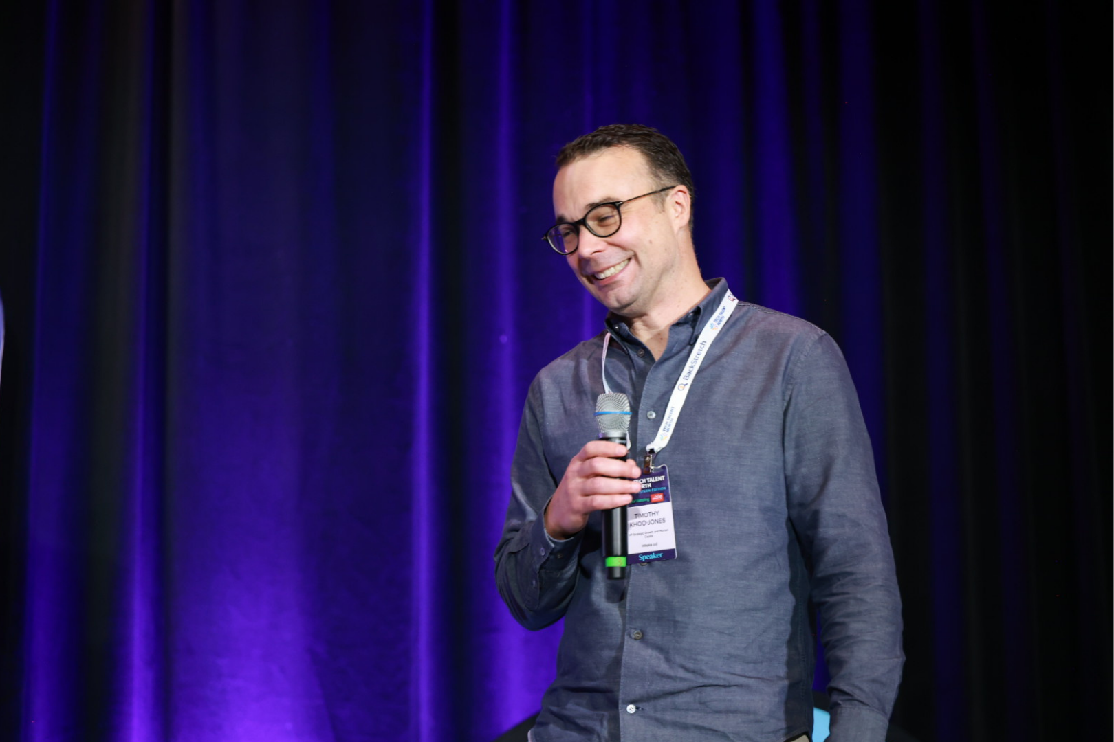 a man on stage, smiling as he presents, holding a microphone