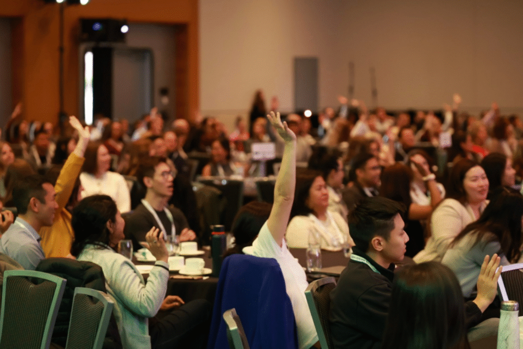 a crowd of people at a keynote address, some of them have their hands raised