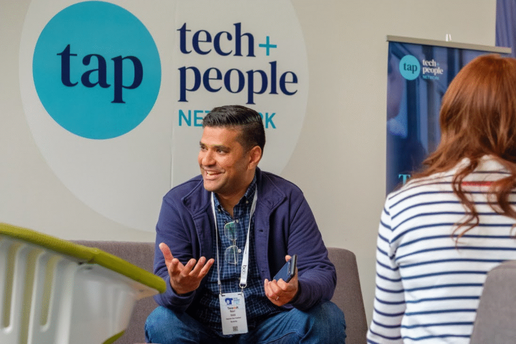 A man sitting down, smiling at someone. A sign on the wall behind him says Tech and People Network