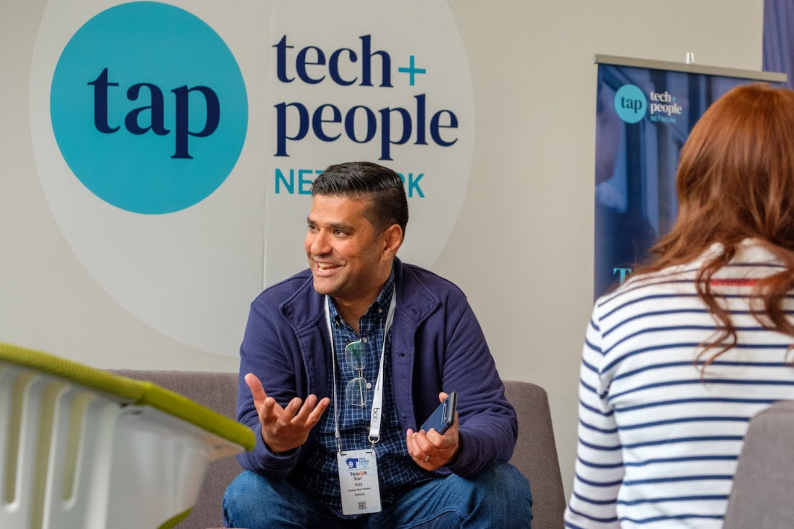 A man sitting down, smiling at someone. A sign on the wall behind him says Tech and People Network