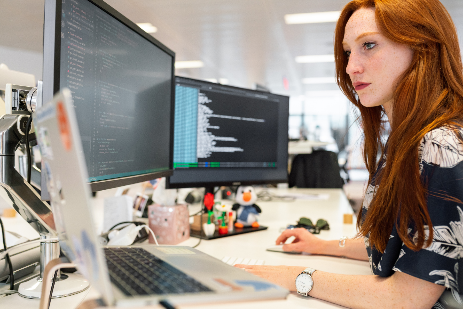 a woman coding software at a desk with 3 monitors.