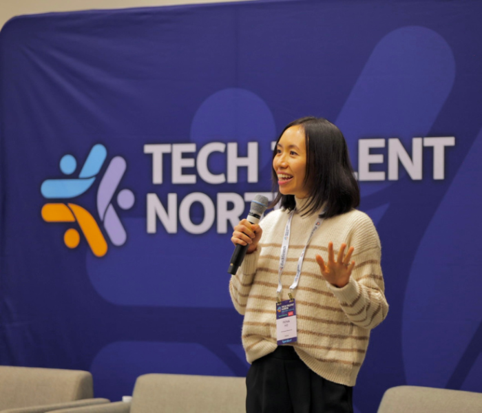 A woman standing in front of a blue banner that says Tech Talent North. She's holding a microphone and smiling as she gives a presentation.